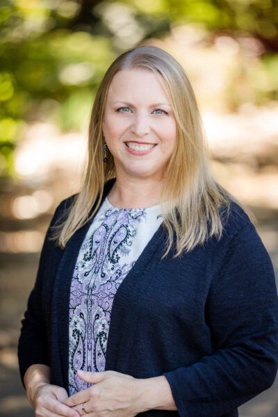 Laura McPhail. White woman with blonde hair wearing a patterned white dress and black cardigan.