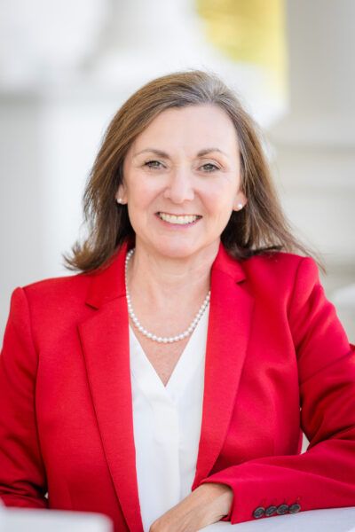 Annette Alexander. White woman with brown hair wearing a white shirt, red blazer, and pearls.